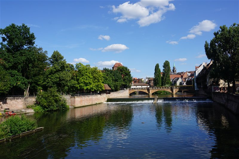 A stone bridge over a river and low weir, framed by trees and old town buildings