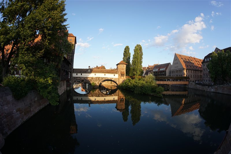 An arched covered bridge and riverside buildings reflected in still water in evening light